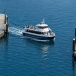 A passenger ferry navigates through a narrow channel between a pedestrian walkway and a dock lined with moored sailboats.