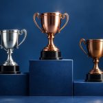 Three championship trophies, one silver and two bronze, are displayed on a blue stepped podium against a dark blue background.